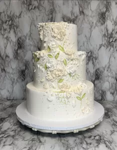 3 tier ivory wedding cake with intricate pale pink florals piped on the side with buttercream with pale green leaves.